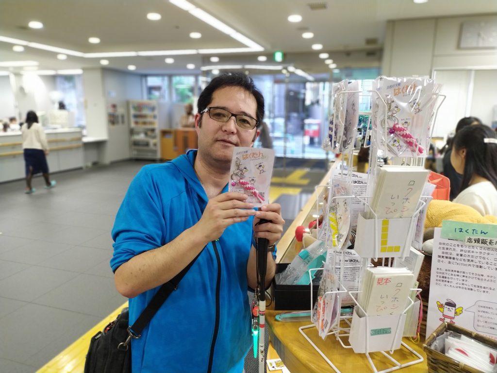 はくたんストラップの商品ラックの隣でストラップを手に持つ渡辺敏之さん