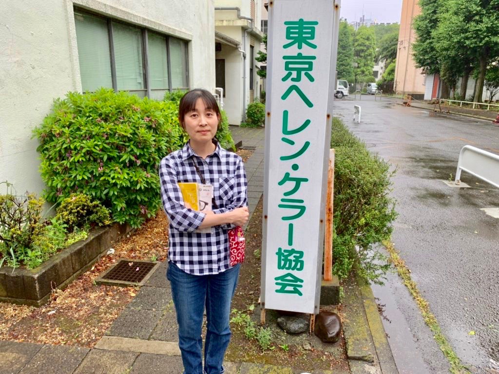 東京ヘレン・ケラー協会の看板の横に立つ小倉さん