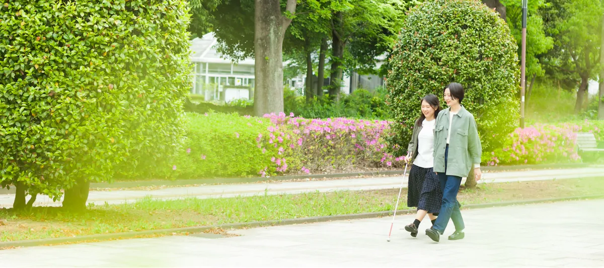 若い女性のヘルパーと視覚障害者が、公園の中を笑顔で歩いているイメージ画像