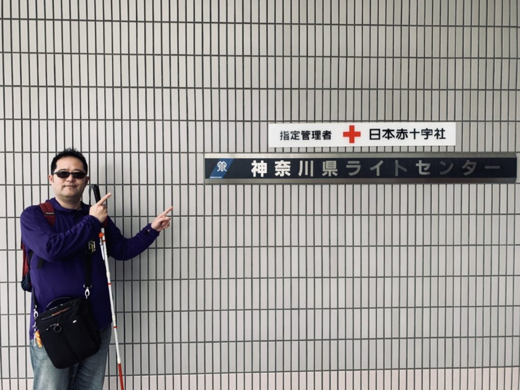 神奈川県ライトセンターの入り口にある看板を指さす渡辺敏之さん