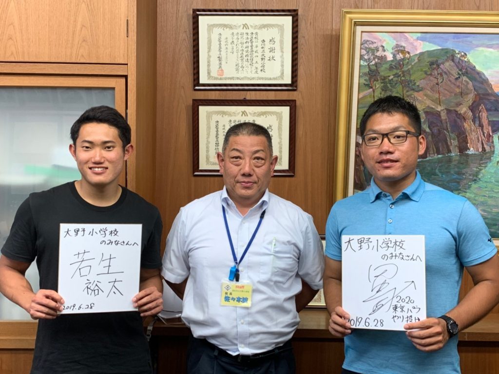 校長室で、若生くんと田中さんがサイン色紙を持ち、校長先生と記念撮影をしている画像