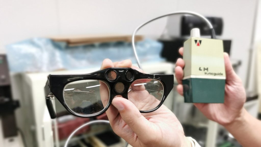 手の平サイズの四角いケースからコードでつながった眼鏡(ソニックガイド)を手に持って撮影した画像。