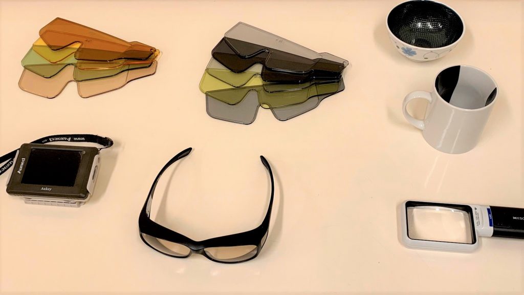 遮光眼鏡やルーペ、中身が見やすいマグカップなどの便利グッズを机の上に並べている画像。