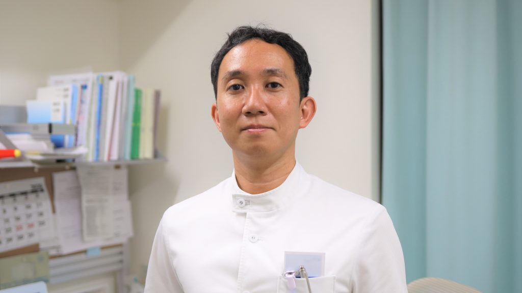 診察室で立っている翁長先生の上半身を撮影した画像。