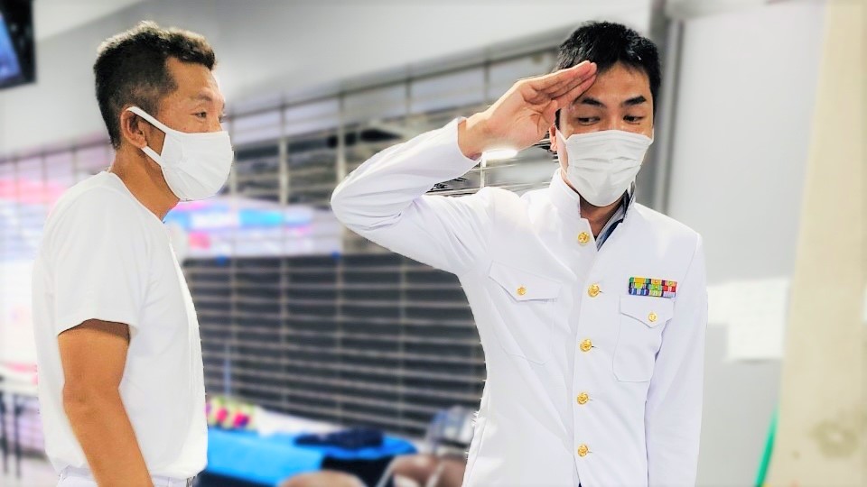 海上自衛隊の白い制服を着て、敬礼のポーズをしている村松さんの画像。