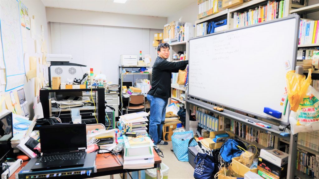 研究室の中で元木さんが立ったまま書棚に手を伸ばして、こちらを向いている画像。