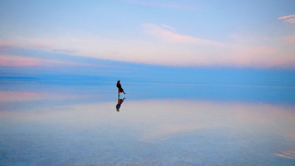 ウユニ塩湖に空の青い色が反射している幻想的な風景の画像。中心に人が一人小さく写っている。