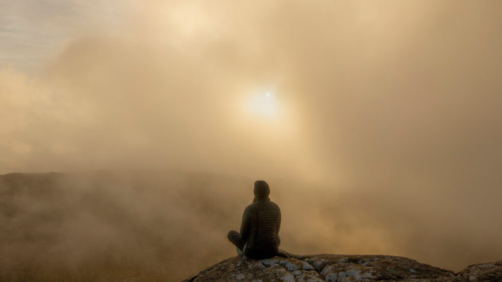 モヤのかかる岩山の頂上で、ぼんやりとした朝日を眺める人