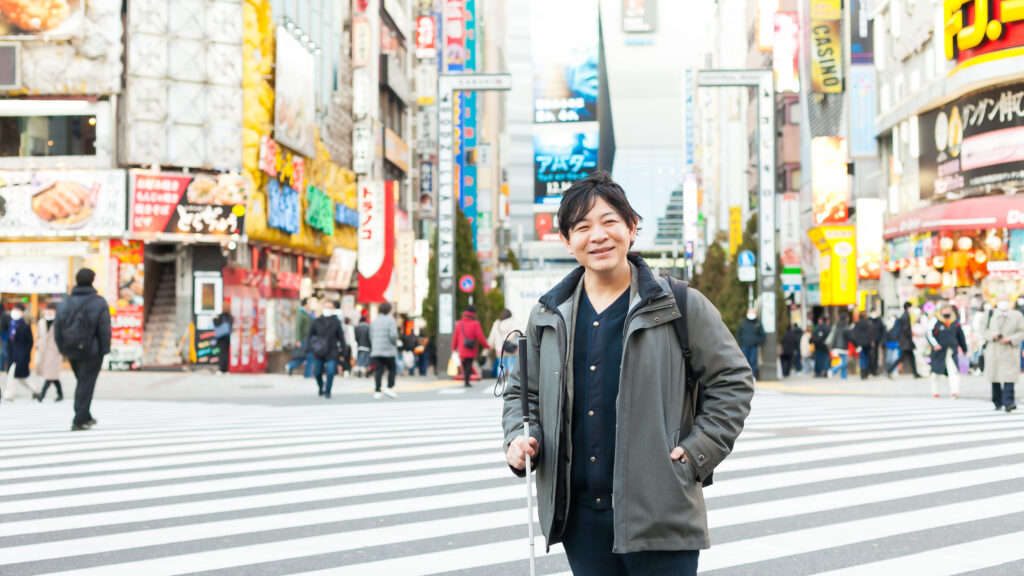渋谷のスクランブル交差点前で、白杖を手に微笑む西川さん。