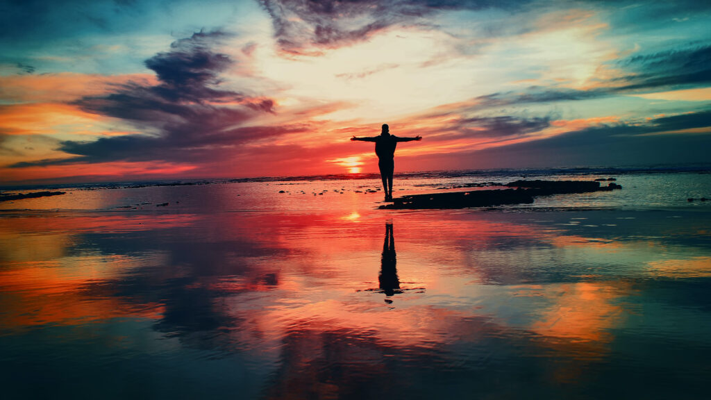 夜明けの時間、鏡のように反射する湖の上に手を広げた人が立っている写真。