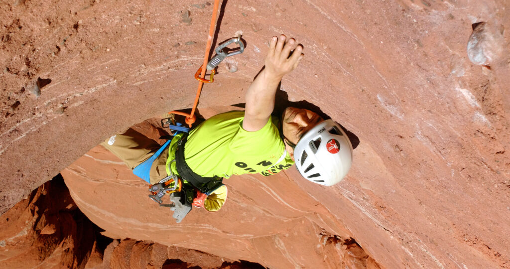 絶壁の赤い岩肌で左手を伸ばしている小林さんの写真。