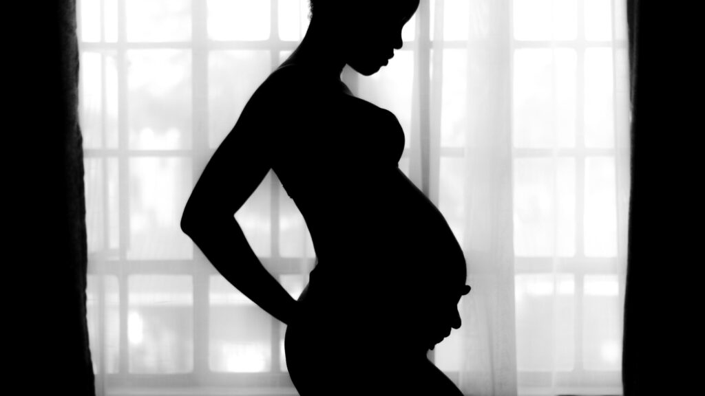 窓際に立つ妊婦。差し込む光に照らされて、妊婦の影が目立つモノクロ写真。
