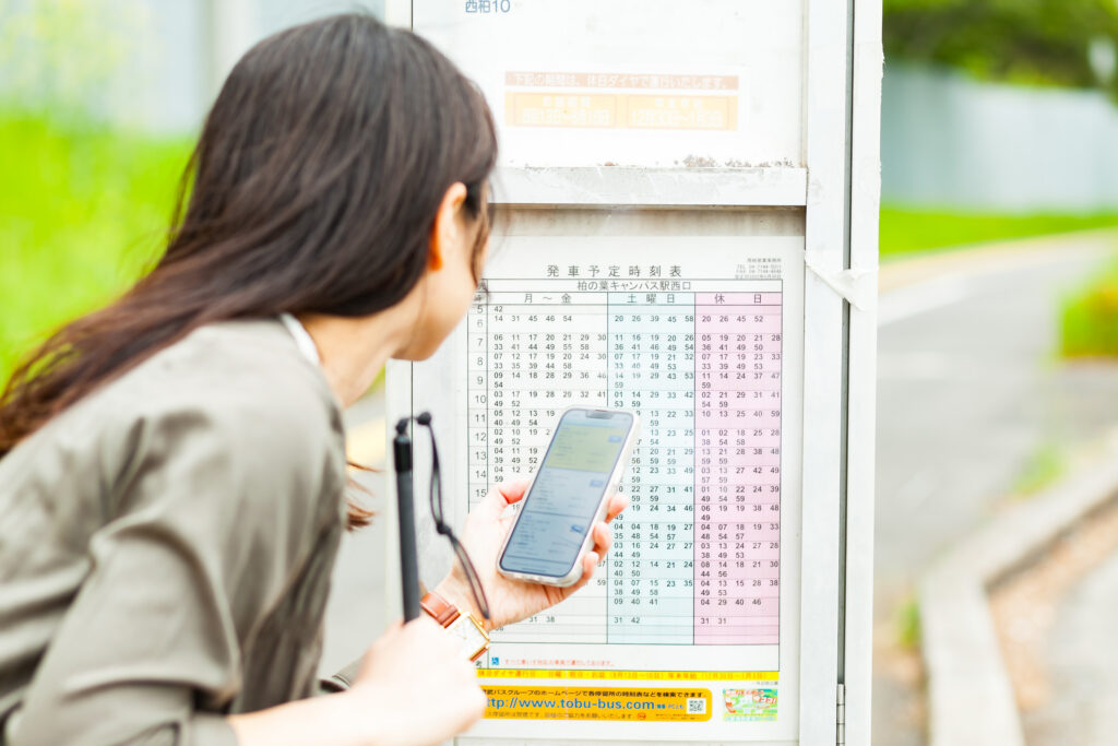 スマートフォンを使いながらバスの時刻表を確認している白杖の女性。