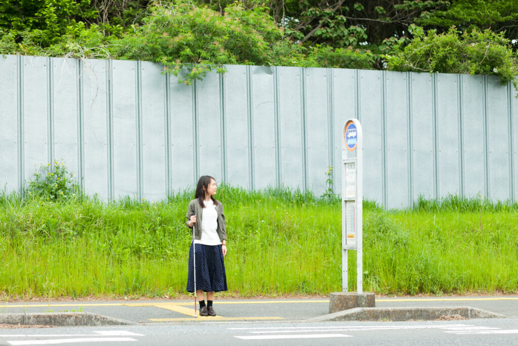 白杖を持った人がバス停でバスを待っている写真。