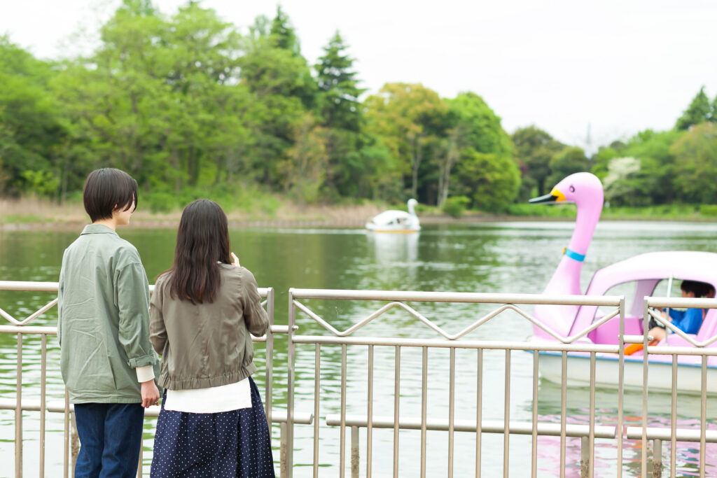 公園の池のほとりで、ピンク色のスワンボートを眺める、白杖の人とガイドヘルパー。