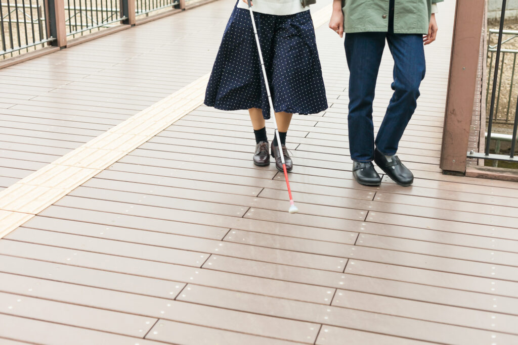 点字ブロックの横を歩く、白杖を持った視覚障害者とガイドヘルパーの足元の写真。