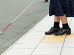 点字ブロックの上で立ち止まる白杖の人の足元の写真。