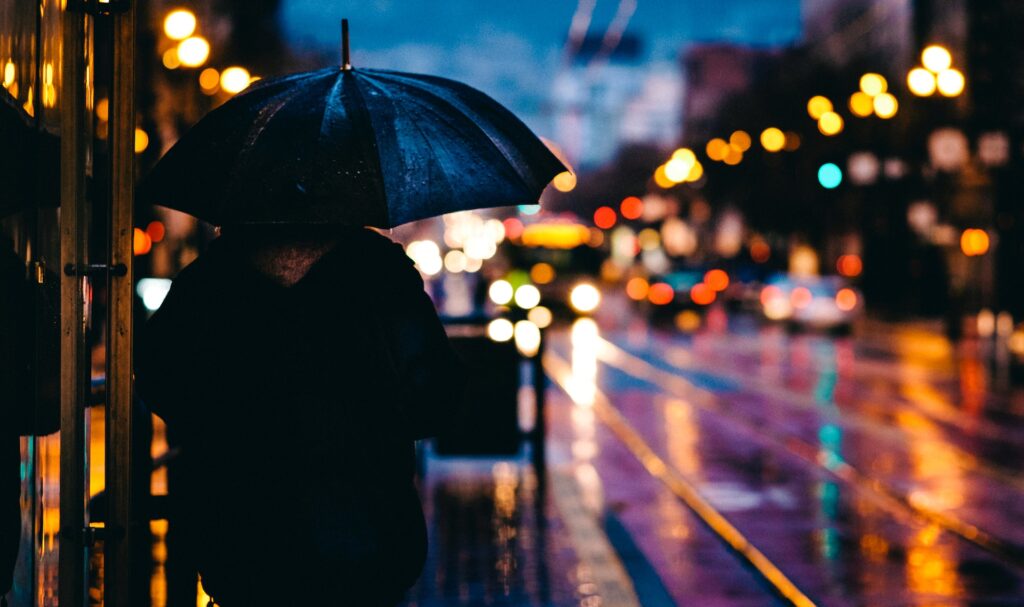 雨が降った夜の街。車や建物の照明がにじんでいて、傘を差した人が立っている。