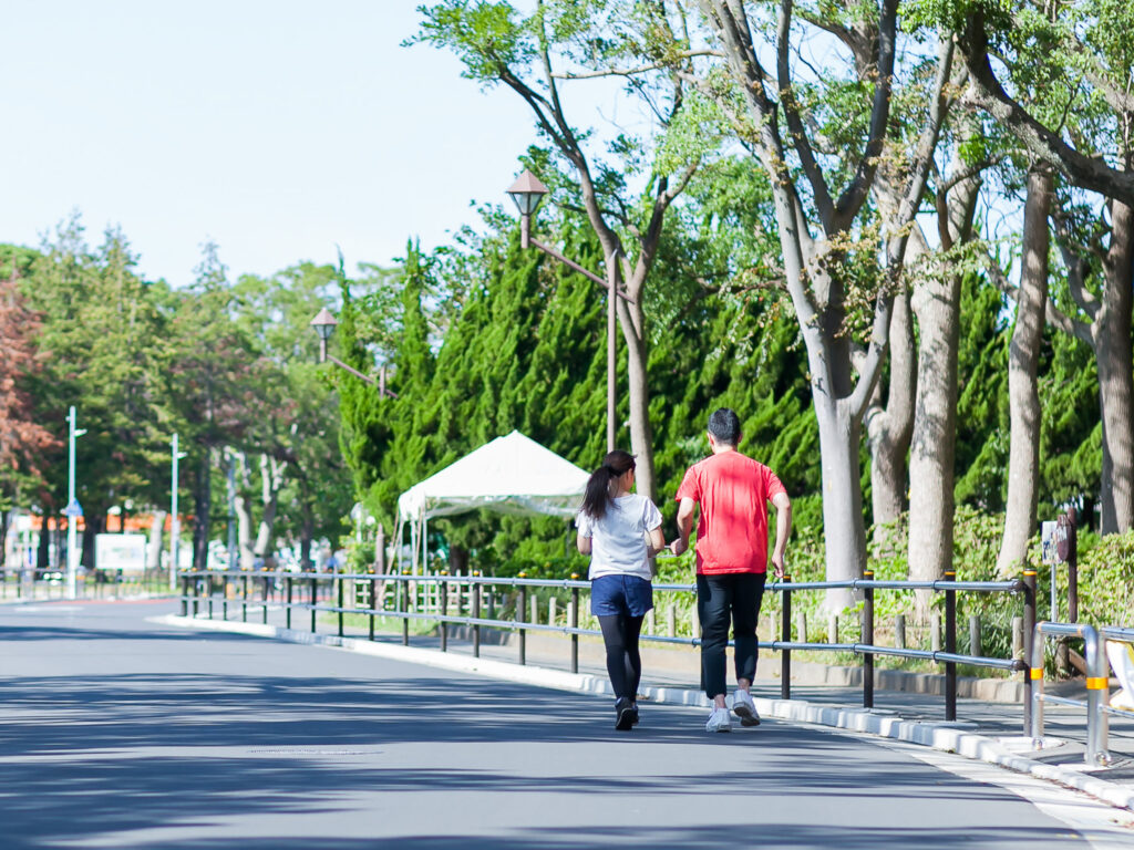 公園をランニングする視覚障害者の女性とタスキを持って伴走する男性の後ろ姿。
