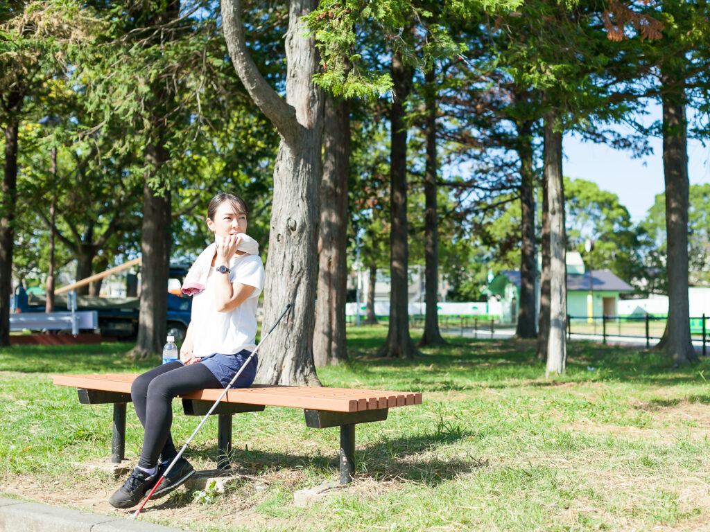 公園のベンチに座る運動着姿の視覚障害者。白杖とペットボトルが隣に置き、タオルで顔を拭いているところ。