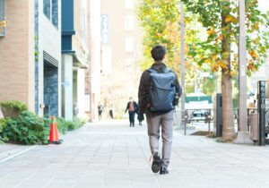 白杖を持った人が、リュックを背負ってオフィス街を歩いている後ろ姿。