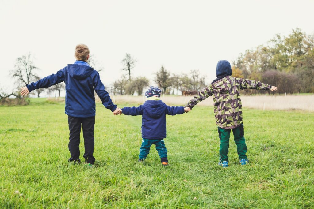 冬服を着て、芝生の上で手をつなぐ三人の子どもの後ろ姿。左から8歳くらい、2歳くらい、5歳くらいの子ども。