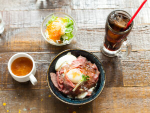 テーブルの上のランチセットの写真。中央にローストビーフ丼、9時の位置にスープ、11時の位置にサラダ、1時の位置にアイスコーヒーがある。