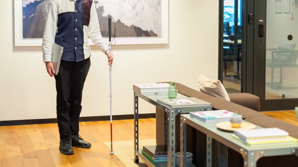 白杖とノートパソコンを持って、オフィス内に立っている男性。
