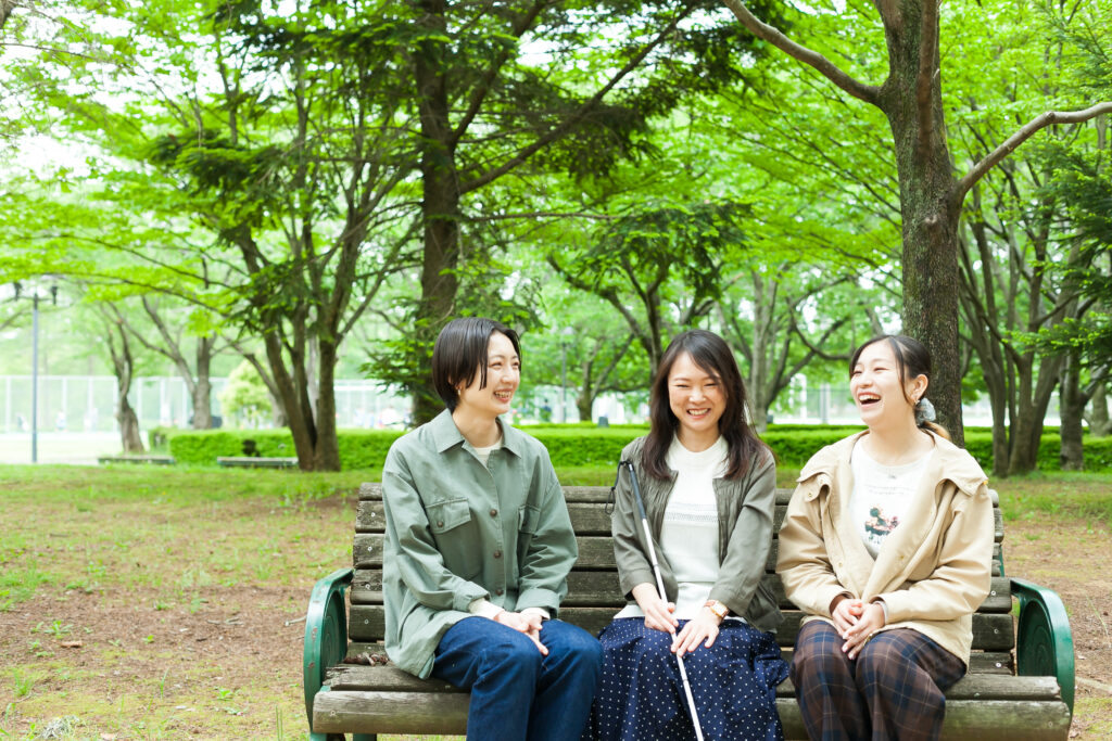 視覚障害者の女性と晴眼者の女性二人が、公園のベンチに座って楽しそうに笑いながら話している。
