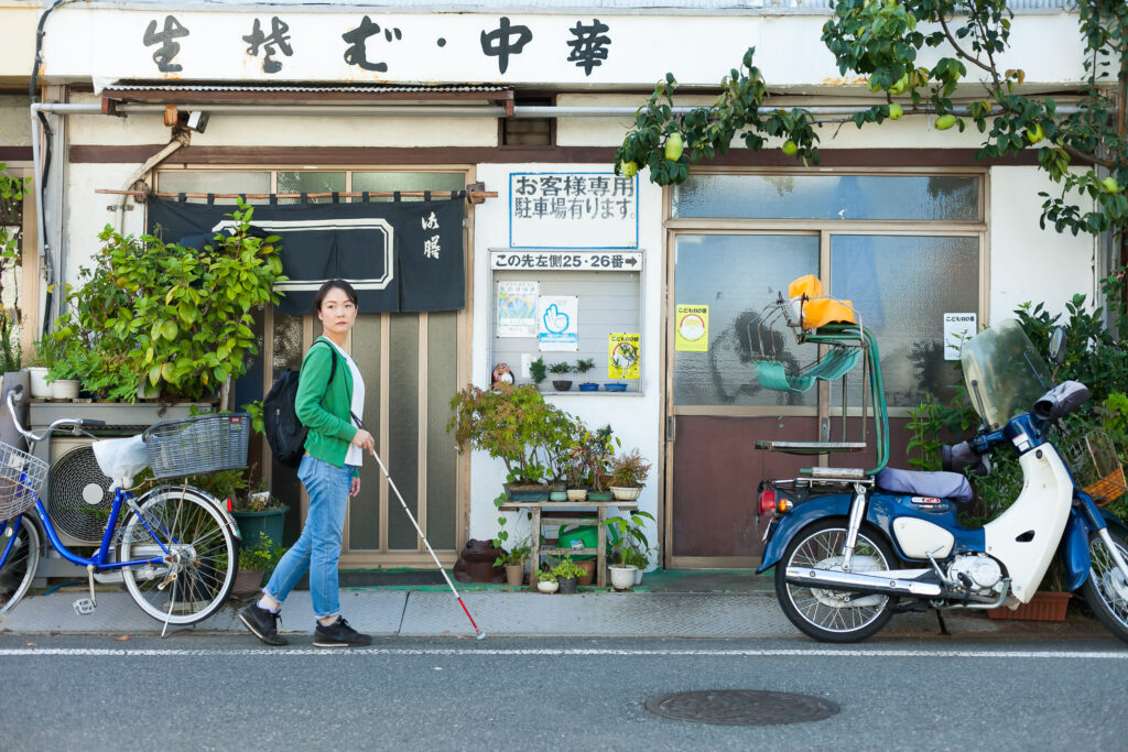 町の中を歩く白杖の女性。蕎麦屋があり、その前に自転車やバイクが止まっている。