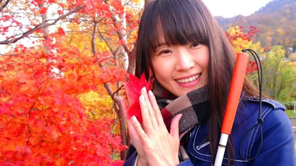 真っ赤な紅葉の中で、青いコートを着て白杖をもって微笑む杉本梢さん。