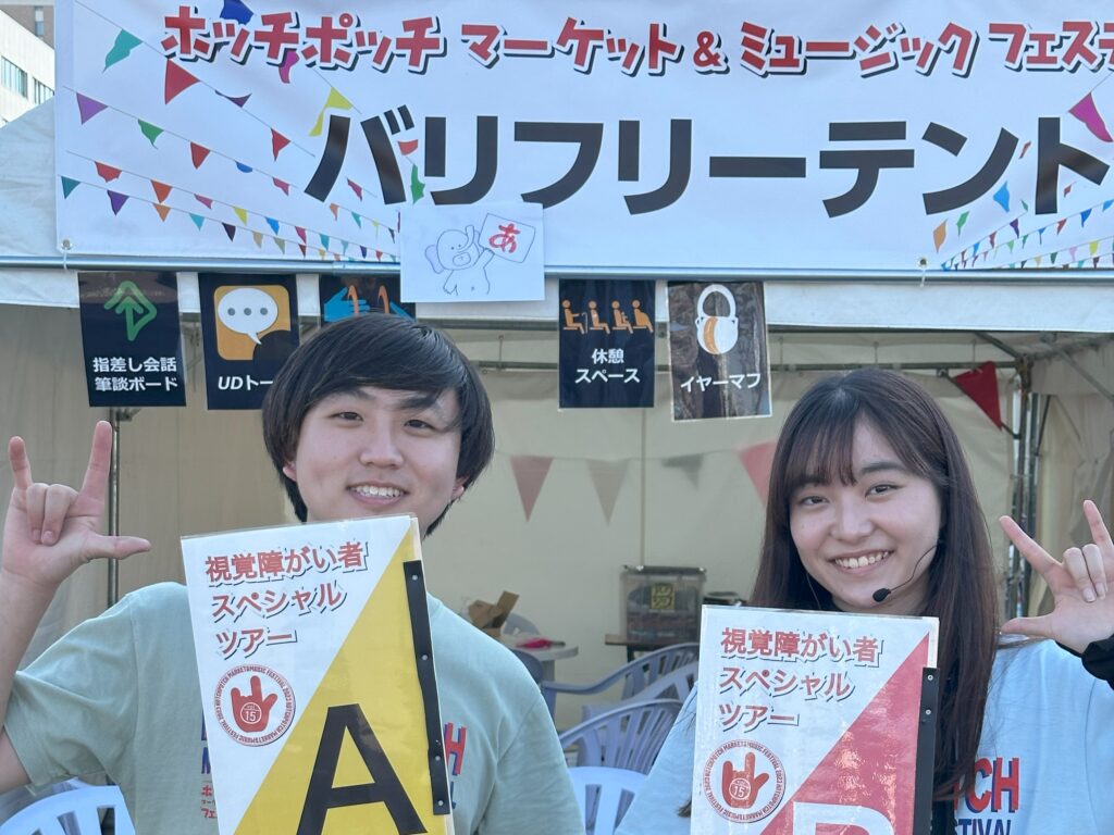 バリアフリーテントの表示があるテントの前で、視覚障害者スペシャルツアーのプラカードを持った笑顔の大野さんと藤原さん。