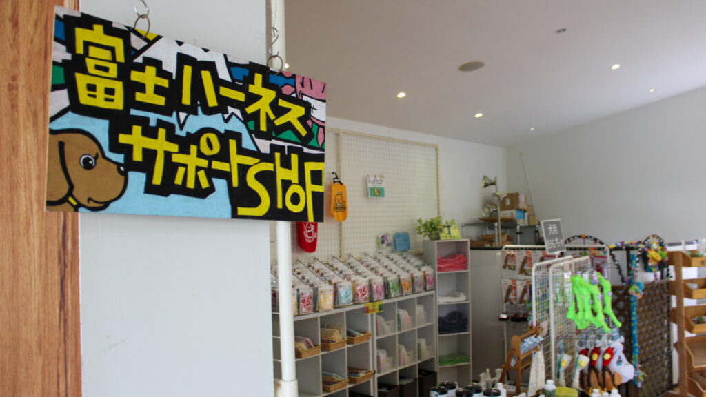 手書き風デザインで「富士ハーネスサポートショップ」と書かれた看板と、店内のグッズの様子。