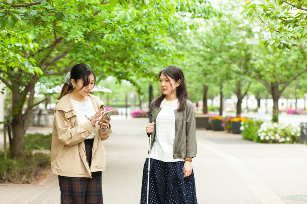 公園の広い歩道の脇に、視覚障害者とガイドヘルパーが立っている。ガイドヘルパーがスマホを操作している。