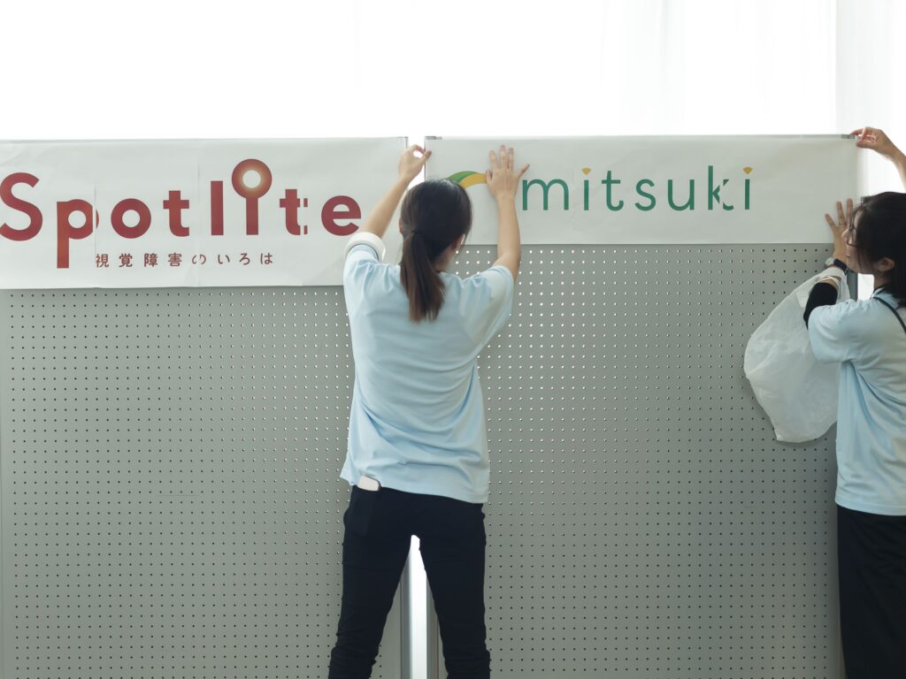 イベント準備の様子。パーティションに「Spolite」と「mitsuki」のロゴマークをプリントした用紙を貼っているスタッフ。