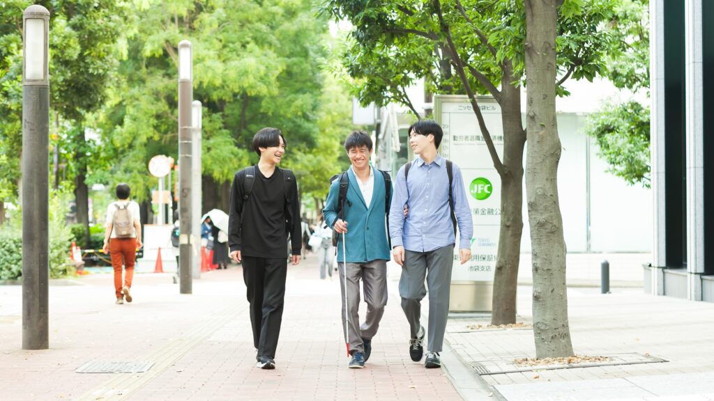 都心の並木道を白杖を持った村竹さんと晴眼者の男性が、三人で談笑しながら歩いている。