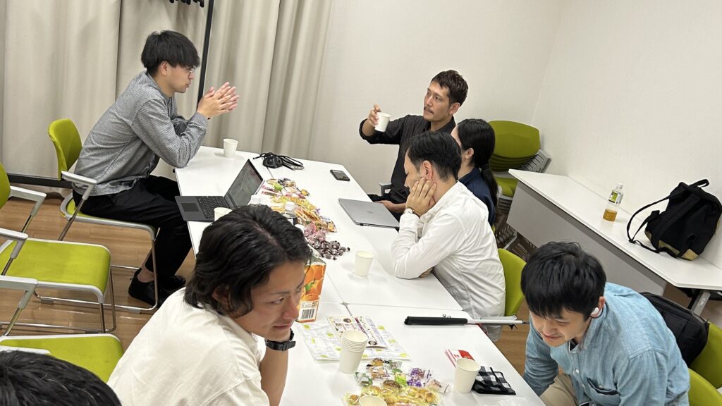 長机に、飲み物やお菓子が並べられ、その周りで視覚障害者やヘルパーが談笑している様子。