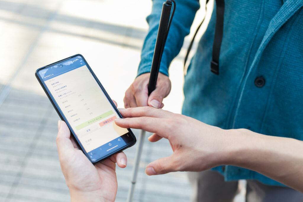 屋外で、白杖の人と、スマホを持っている人の手元。スマホの画面にはおでかけくんが表示されている。