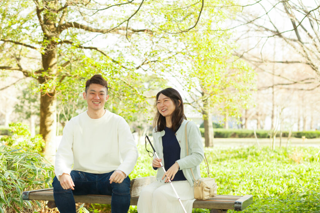 緑の多い公園のベンチに座って、笑顔で話している白杖の人と高橋さん。