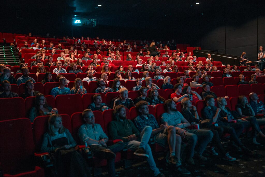 映画館の客席のイメージ写真。暗い大きな室内に赤い客席が並び、たくさんの人が座っている。