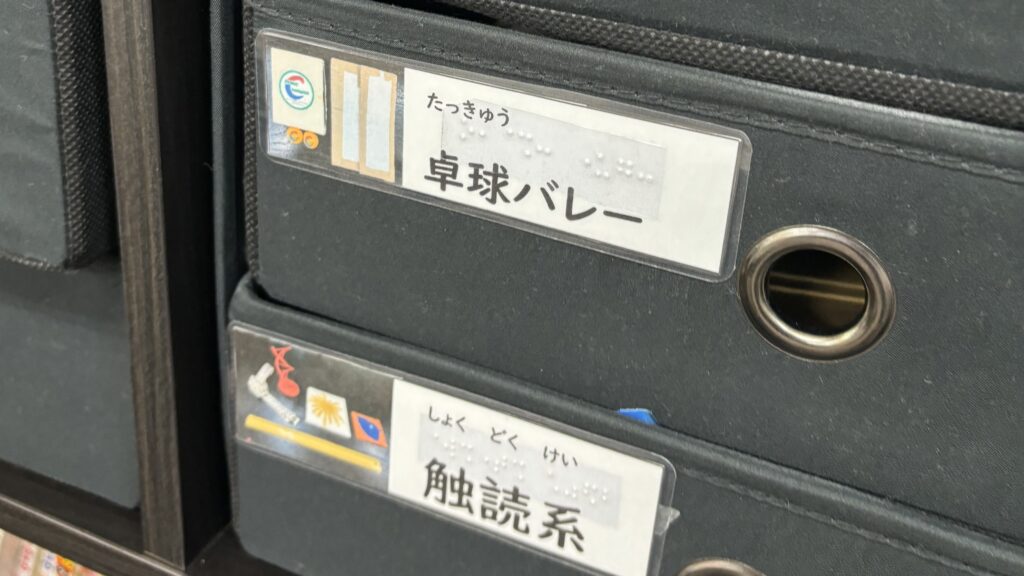 療育に使う道具類をしまっている引き出し。「卓球・バレー」「触読系」などの墨字表記と点字表記、道具類の写真がついたラベルが貼ってある。