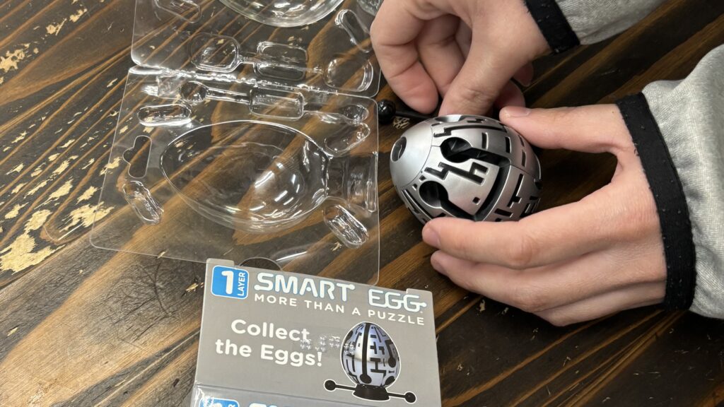 卵の形をした銀色の立体パズル。「SMART  EGG
 MORE THAN APUZZLE」「Collect the Eggs!」と書かれた台紙があり、点字もつけられている。