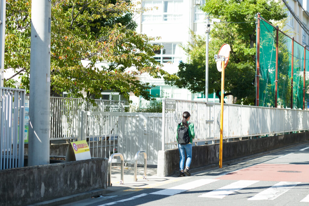 学校らしき建物の横を歩いている、グリーンのカーディガン姿の白杖の人。