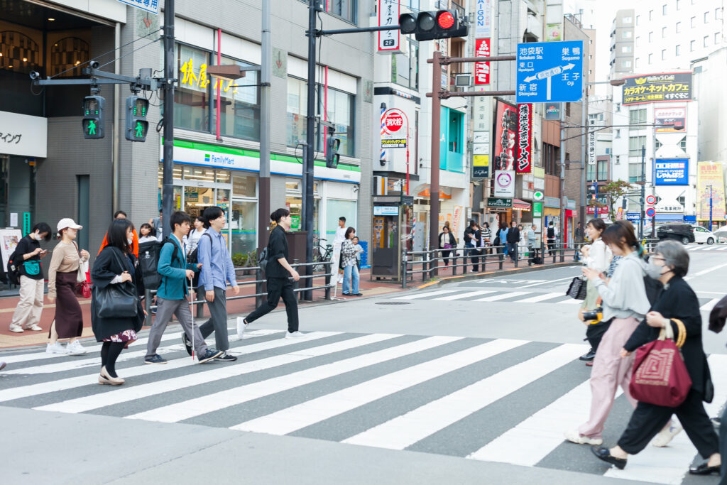 人通りが多くにぎやかな大通り。人が行き交う交差点で横断歩道を白杖の人とガイドヘルパーがわたっている。