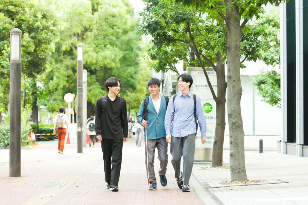 緑が生い茂る路上で、横に並んで歩く3人の笑顔の男性たち。真ん中の男性は右手に白杖を持ち、左手で左隣の男性の腕を借りて歩いている。