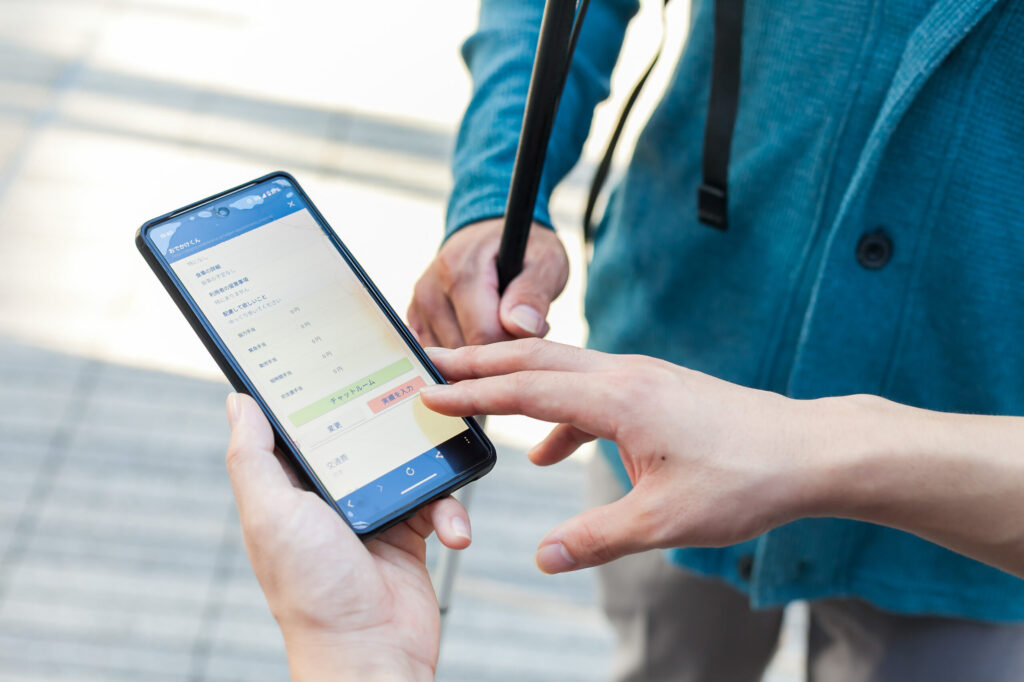 スマートフォンで「おでかけくん」を操作する男性の手。背景には白杖を持つ別の男性の右腕が映っている。