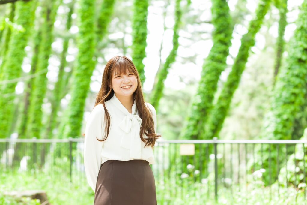 緑色の木々を背景に、白いブラウス姿で笑顔で立っているガイドヘルパーのあだっちゃん。