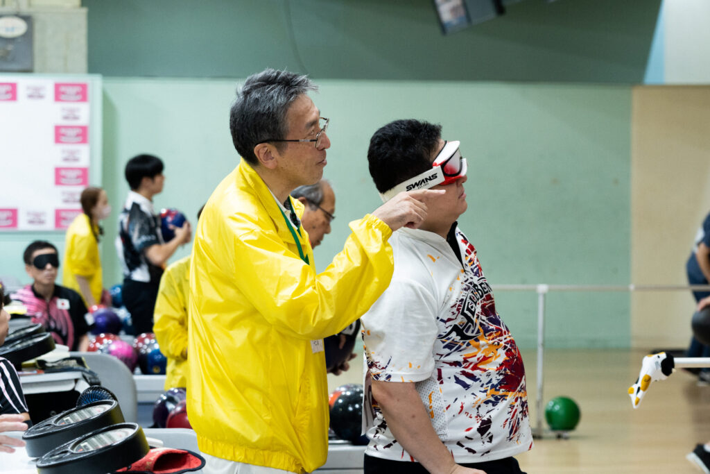 黄色いジャンパーを着たアシスタントに、残ったピンを教えてもらうアイマスクをした井上さん。