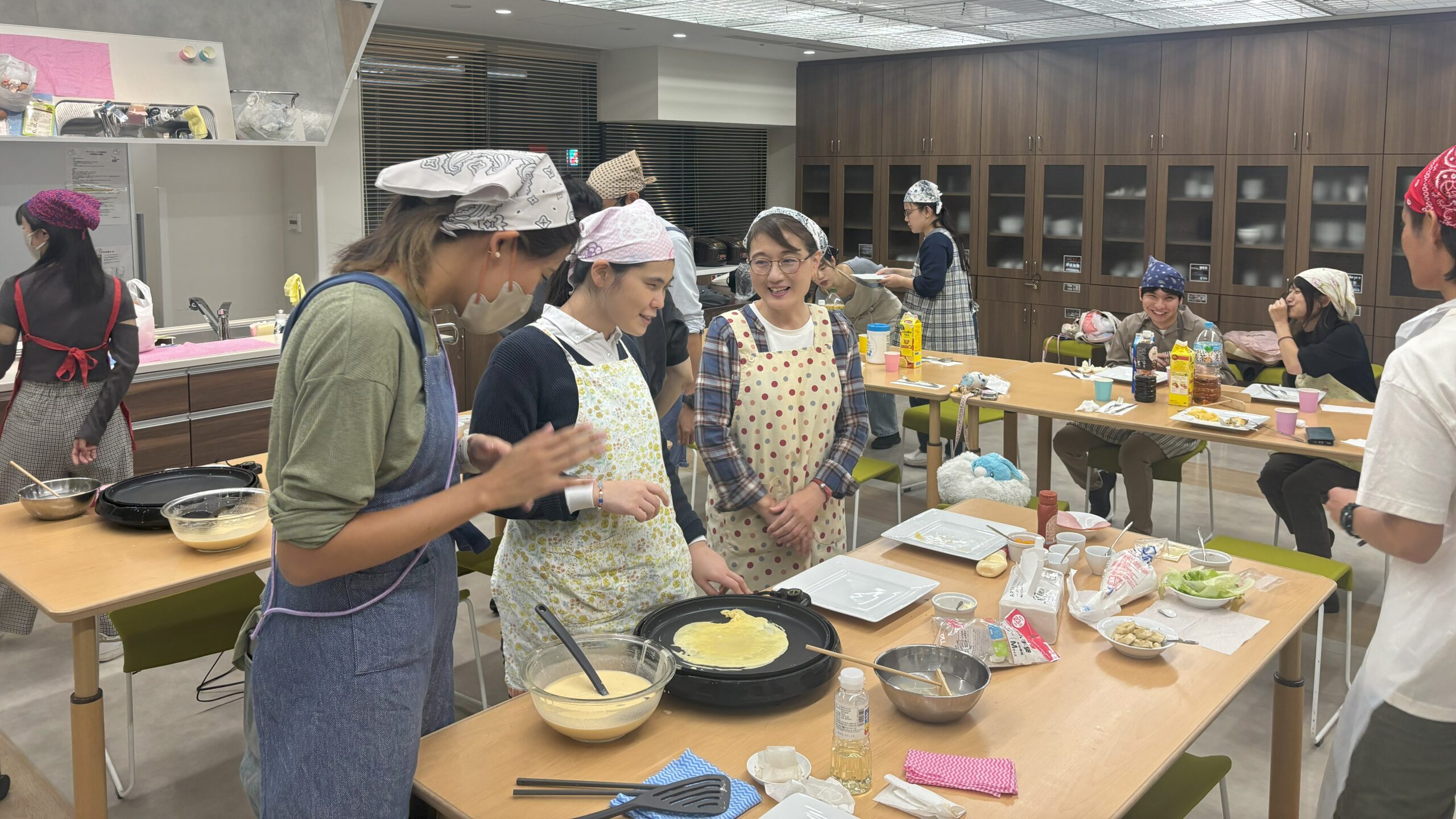 クレープ教室の当日の様子。キッチンのあるレンタルスペースにテーブルが並べられ、2台以上のホットプレートでクレープを焼いているところ。視覚障害者もヘルパーも笑顔で楽しそうな様子。