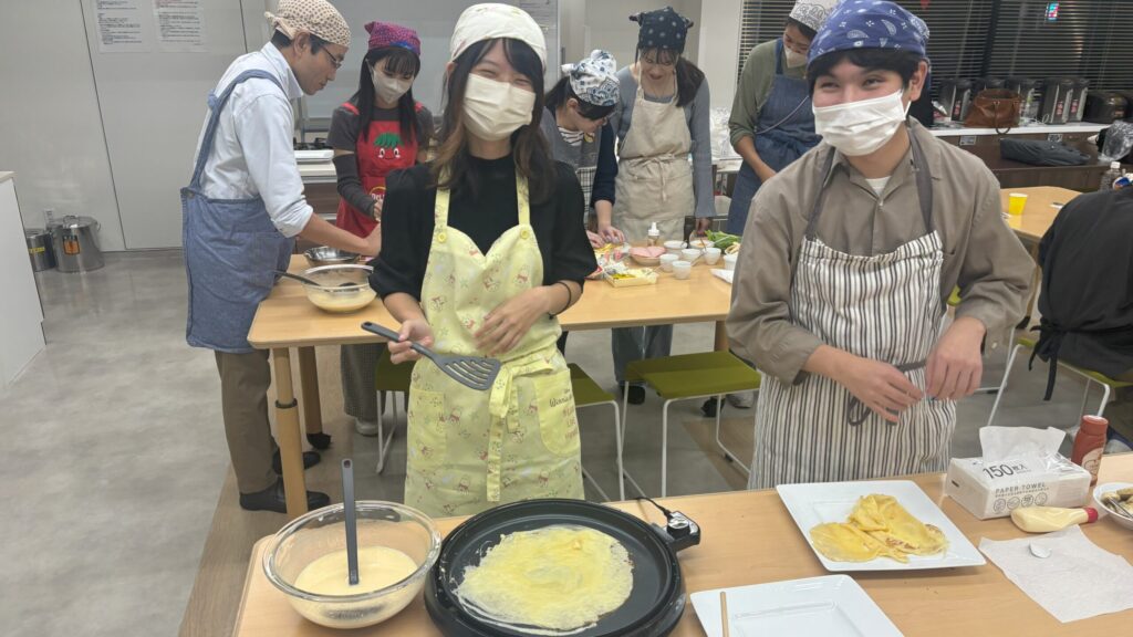 ホットプレートでクレープ生地を焼いている視覚障害者とガイドヘルパー。お皿の上に焼きあがったクレープが畳んで盛り付けられている。